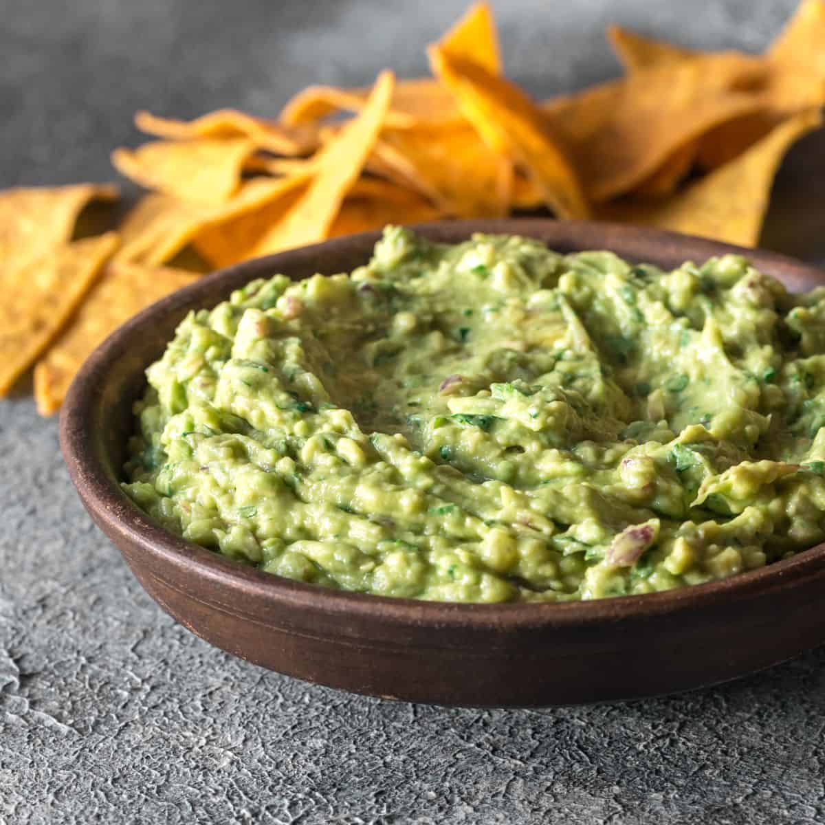 Guacamole in a brown bowl with chips in the background.