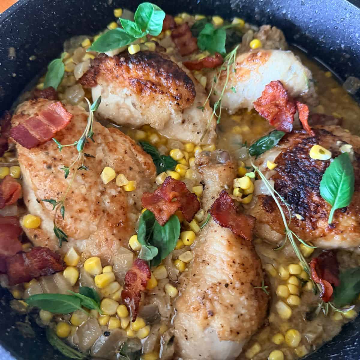 Pan-seared chicken with golden-brown crusts in a skillet with corn, crispy bacon pieces, caramelized onions, and fresh herbs including basil and thyme in a cream sauce