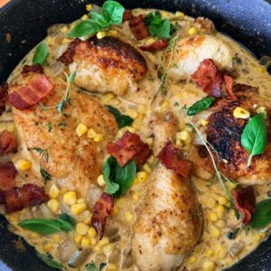 A cast iron skillet filled with golden-brown seared chicken thighs arranged in a circle over creamy corn sauce, garnished with crispy bacon pieces, fresh basil leaves, and delicate thyme sprigs, photographed from above with an orange striped kitchen towel visible at the edge.