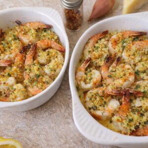 Two small white baking dishes filled with baked shrimp scampi, topped with a golden breadcrumb mixture and herbs, with lemon slices and pantry ingredients nearby on a light countertop.
