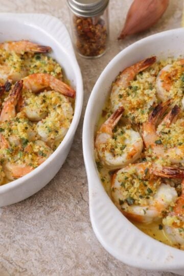 Two small white baking dishes filled with baked shrimp scampi, topped with a golden breadcrumb mixture and herbs, with lemon slices and pantry ingredients nearby on a light countertop.