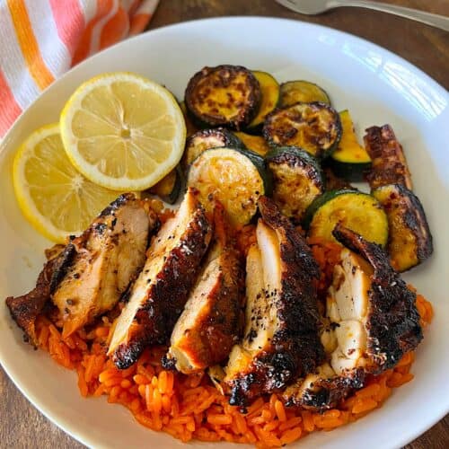 White plate with sliced grilled chicken breast served over orange Spanish rice, alongside charred zucchini rounds and two fresh lemon slices, photographed from above with an orange striped kitchen towel visible at the edge.