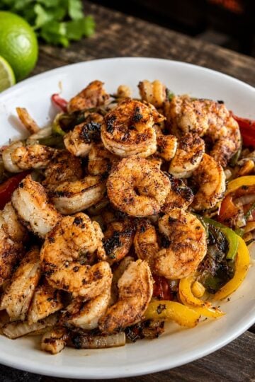 Seasoned shrimp fajitas served in a white bowl with sautéed bell peppers, onions, and tomatoes on a rustic wooden table with fresh lime wedges.
