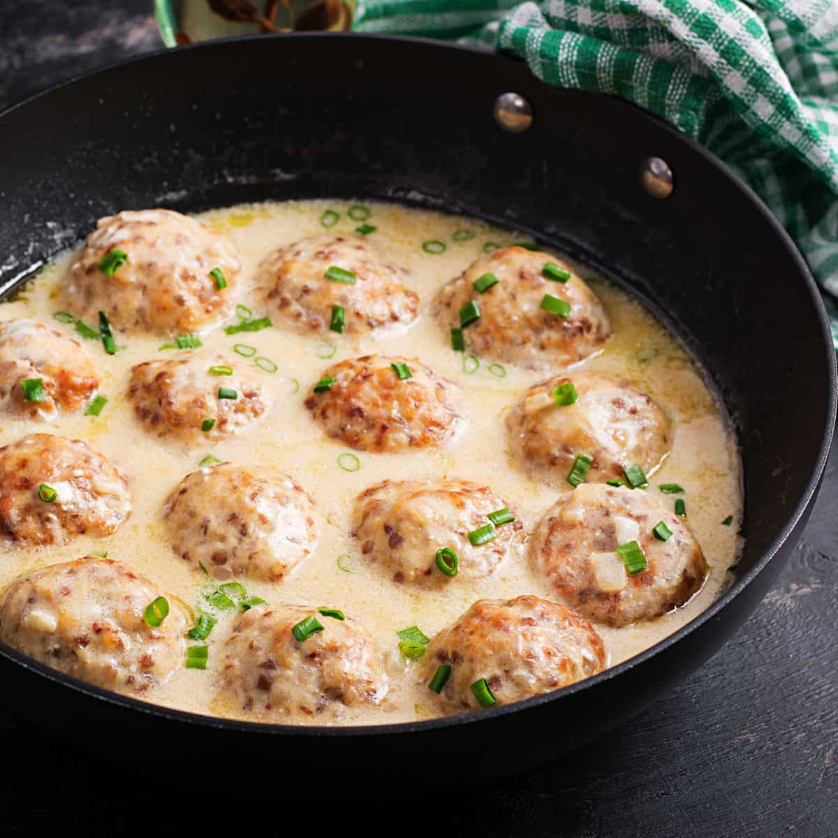 Buffalo chicken meatballs simmering in a creamy sauce, garnished with chopped green onions in a black skillet.