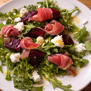 Fresh arugula salad topped with roasted beet slices, rolled prosciutto, and crumbled goat cheese on a white plate, lightly dressed with olive oil.