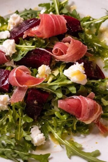 Fresh arugula salad topped with roasted beet slices, rolled prosciutto, and crumbled goat cheese on a white plate, lightly dressed with olive oil.