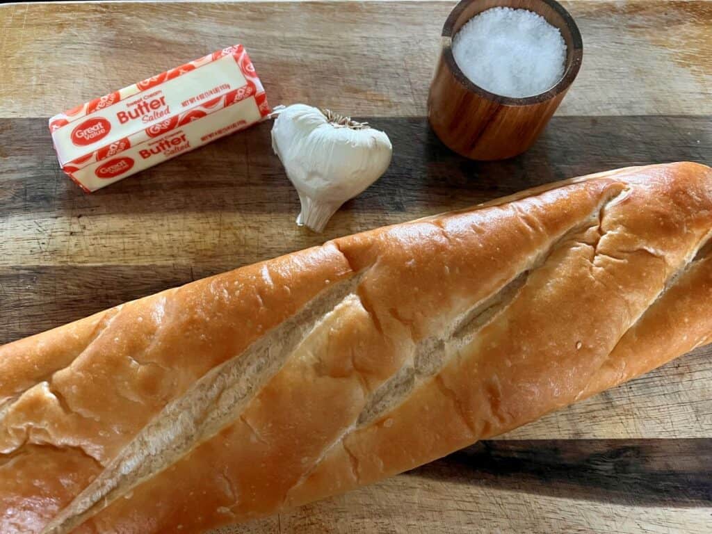 French bread, a head of garlic, a stick of butter and salt on a cutting board