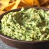 Guacamole in a brown bowl with chips in the background