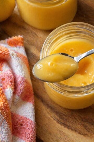 Close-up of a spoon lifting thick, glossy lemon curd from a small glass jar set on a wooden cutting board, with an orange-and-white kitchen towel draped beside it and whole lemons softly blurred in the background.