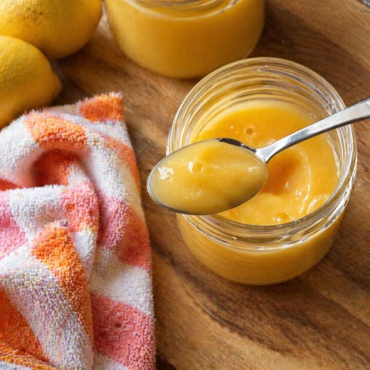 Close-up of a spoon lifting thick, glossy lemon curd from a small glass jar set on a wooden cutting board, with an orange-and-white kitchen towel draped beside it and whole lemons softly blurred in the background.