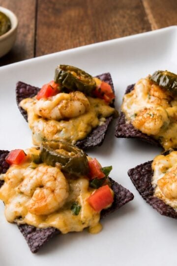White plate with cheesy shrimp nachos topped with melted cheese sauce over blue corn tortilla chips, garnished with diced tomatoes and peppers,