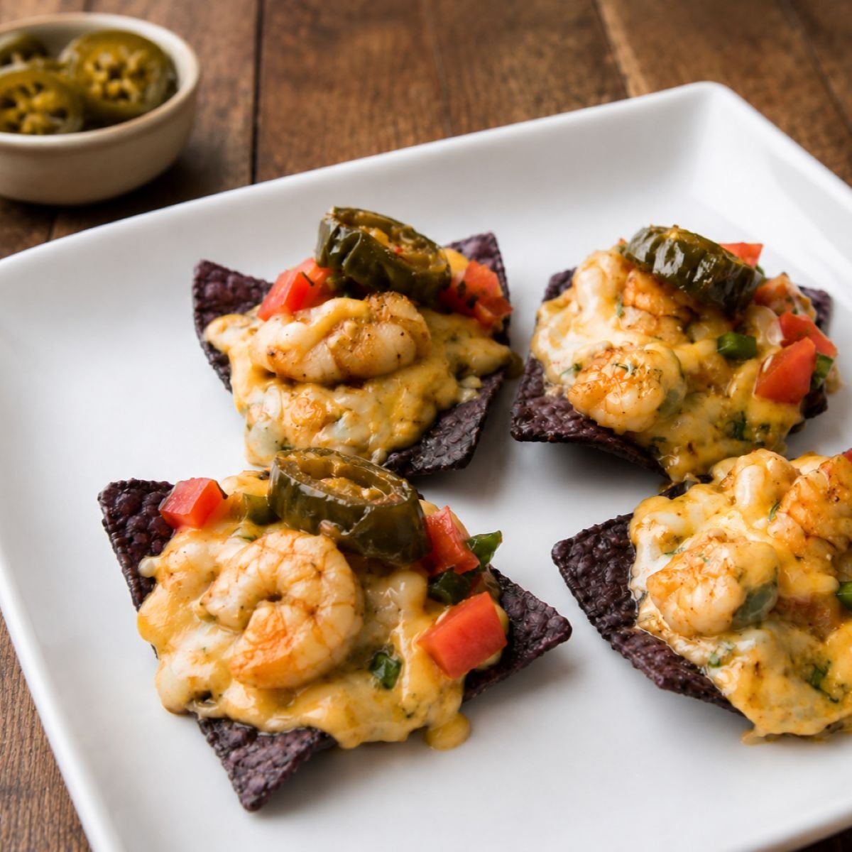 White plate with cheesy shrimp nachos topped with melted cheese sauce over blue corn tortilla chips, garnished with diced tomatoes and peppers,