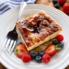 Slice of sopapilla cheesecake on a white plate with a fork drizzled with honey and surounded by strawberries and blueberries.