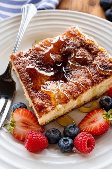 Slice of sopapilla cheesecake on a white plate with a fork drizzled with honey and surounded by strawberries and blueberries.