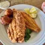 Two pieces of fried catfish on a white plate with hushpuppies and lemon wedges