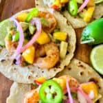 Colorful shrimp tacos on corn tortillas topped with diced mango, sliced jalapeños, pickled red onions, and avocado, garnished with lime wedges on a wooden surface.