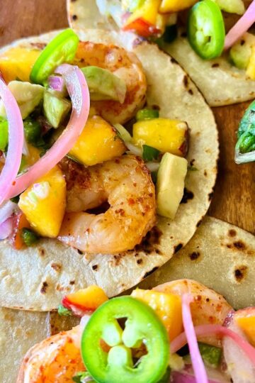 Colorful shrimp tacos on corn tortillas topped with diced mango, sliced jalapeños, pickled red onions, and avocado, garnished with lime wedges on a wooden surface.