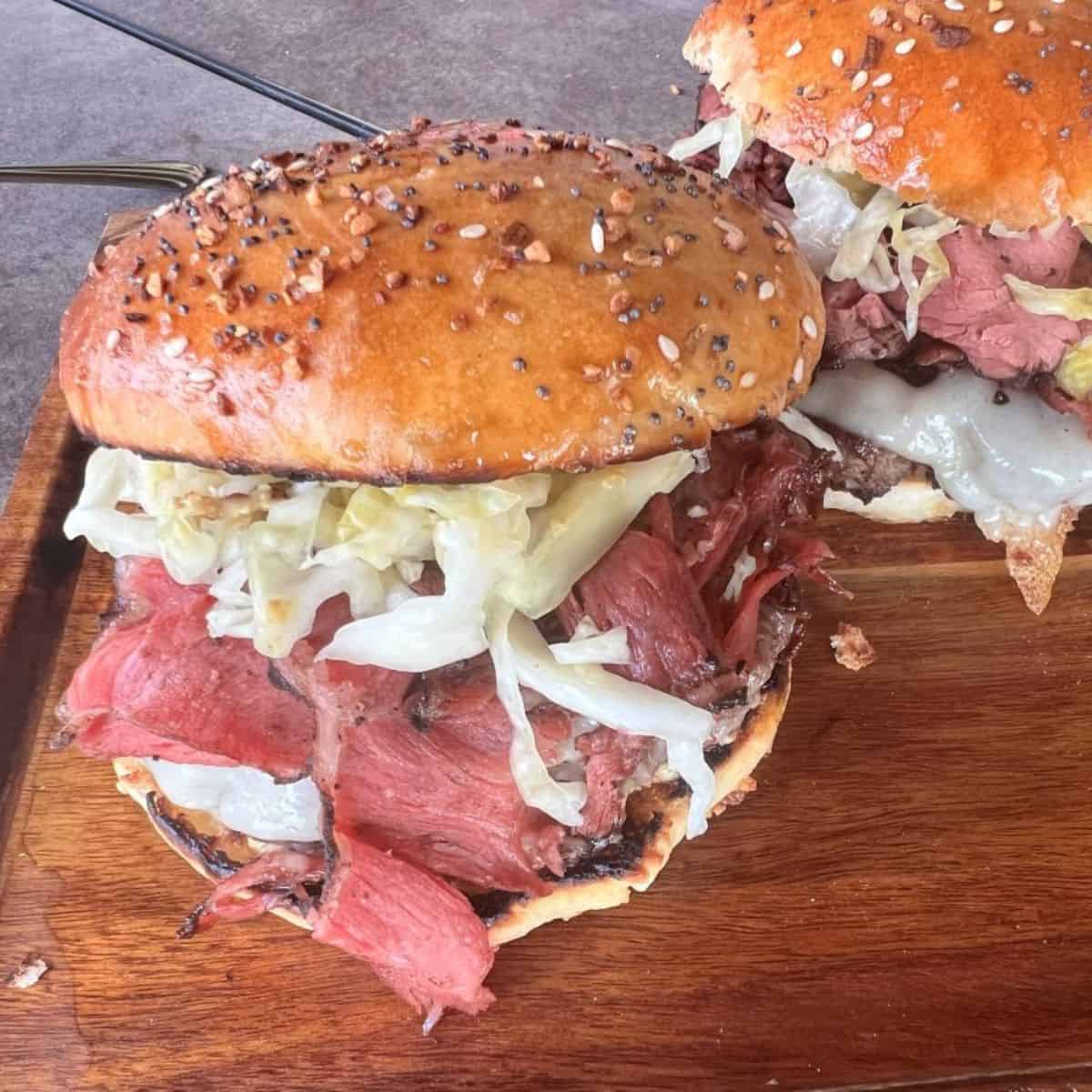 Two fully assembled gourmet burgers on a wooden cutting board, stacked high with beef patties, Swiss cheese, pastrami, and sauerkraut on seeded buns. The burgers appear substantial and ready to eat.