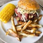 A completed burger meal on a white plate, featuring the layered burger with a seeded bun, accompanied by golden french fries and a corn on the cob. The burger shows all the layers including the meat, cheese, pastrami, and sauerkraut.