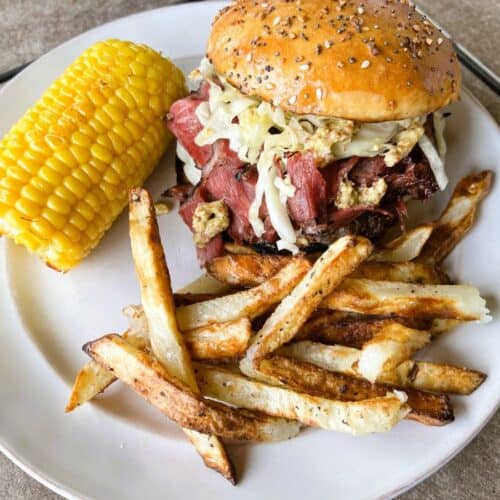 A completed burger meal on a white plate, featuring the layered burger with a seeded bun, accompanied by golden french fries and a corn on the cob. The burger shows all the layers including the meat, cheese, pastrami, and sauerkraut.