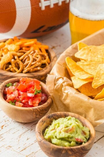 Tortilla chips with guacamole, salsa, and snack mix in wooden bowls, with beer glasses and a football in the background on a white table.