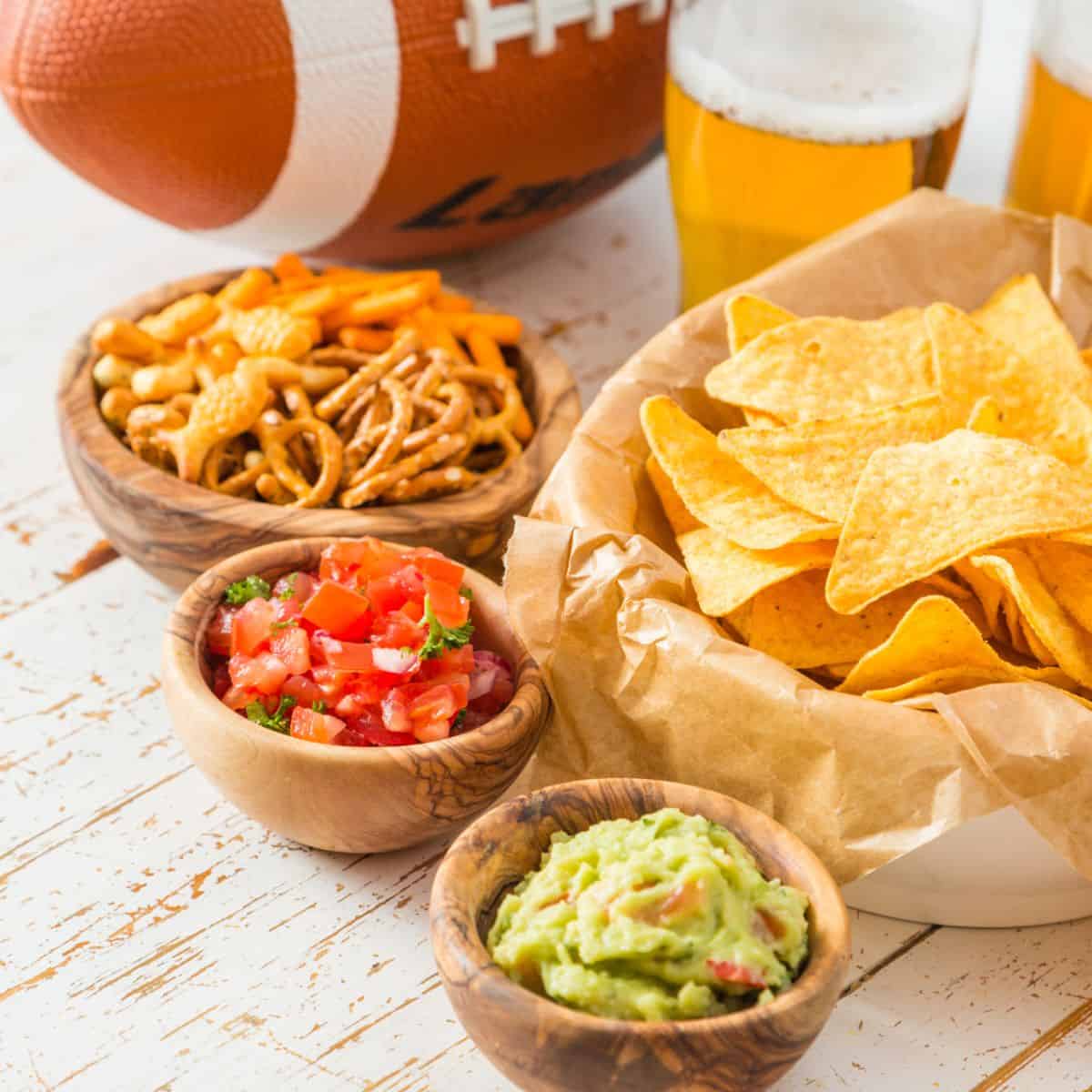 Tortilla chips with guacamole, salsa, and snack mix in wooden bowls, with beer glasses and a football in the background on a white table.