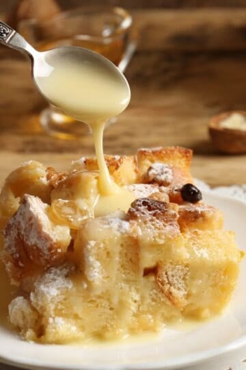 A slicee of bread pudding on a white plate with bourbon cream sauce being drizzled on top
