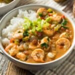 Finished shrimp and chicken gumbo served in a white bowl over white rice, garnished with chopped green onions and red peppers, alongside another bowl of gumbo in the background on a striped cloth napkin.