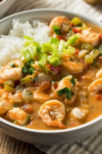 Finished shrimp and chicken gumbo served in a white bowl over white rice, garnished with chopped green onions and red peppers, alongside another bowl of gumbo in the background on a striped cloth napkin.