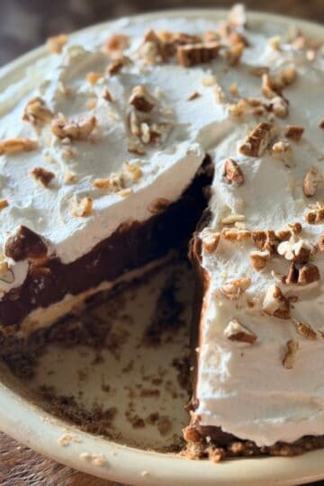 Slice of layered chocolate pie being removed, showing the distinct pecan crust, cream cheese, chocolate pudding, and whipped cream layers with pecan garnish on top.