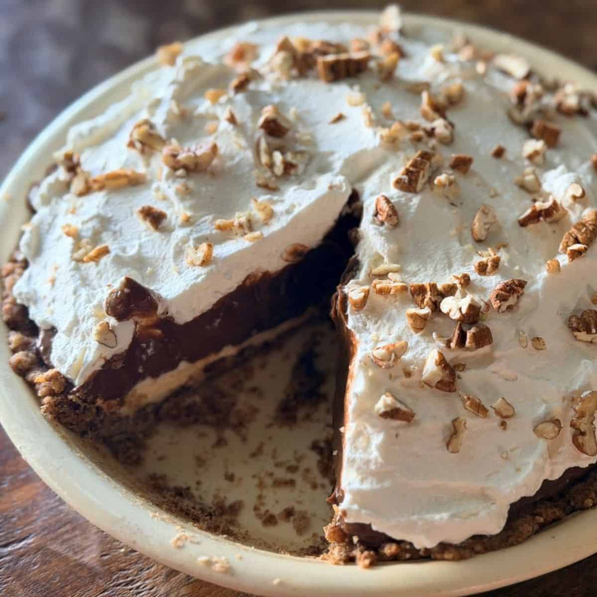 Slice of layered chocolate pie being removed, showing the distinct pecan crust, cream cheese, chocolate pudding, and whipped cream layers with pecan garnish on top.