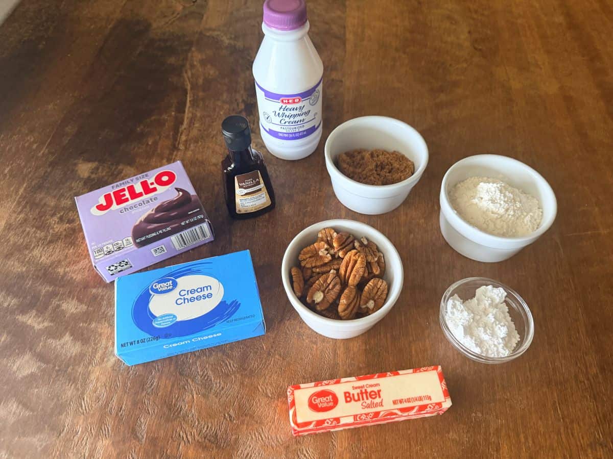 Baking ingredients arranged on a wooden table including a bottle of heavy whipping cream, chocolate Jell-O pudding mix, cream cheese, butter, vanilla extract, bowls containing pecans, brown sugar, flour, and powdered sugar.