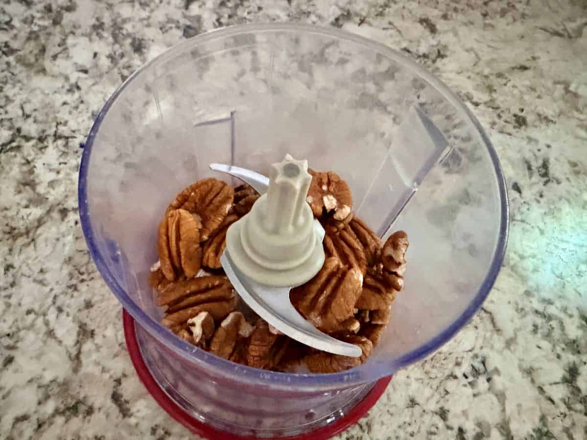 Pecan halves in a food processor bowl with the blade visible, ready to be processed.