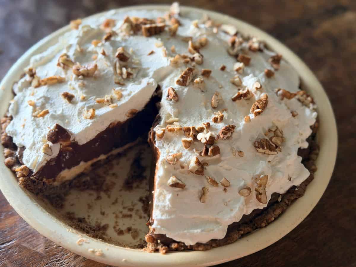Slice of layered chocolate pie being removed, showing the distinct pecan crust, cream cheese, chocolate pudding, and whipped cream layers with pecan garnish on top.