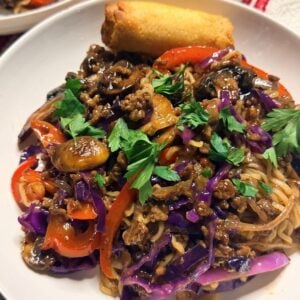 Plated beef ramen noodle dish garnished with fresh cilantro, served alongside an egg roll on a white plate
