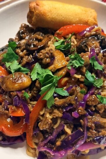 Plated beef ramen noodle dish garnished with fresh cilantro, served alongside an egg roll on a white plate