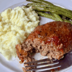 Classic meatloaf dinner plate with a slice of glazed meatloaf, mashed potatoes, and roasted green beans, with a fork taking a bite.