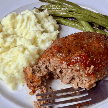 Classic meatloaf dinner plate with a slice of glazed meatloaf, mashed potatoes, and roasted green beans, with a fork taking a bite.