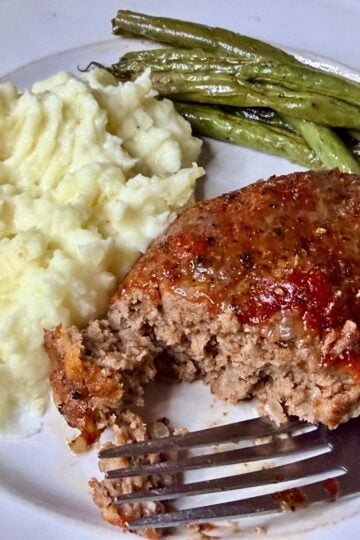 A plated dinner featuring a slice of meatloaf with ketchup glaze, creamy mashed potatoes, and roasted green beans, with a fork on the plate.