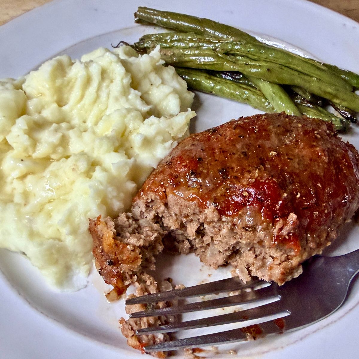 A plated dinner featuring a slice of meatloaf with ketchup glaze, creamy mashed potatoes, and roasted green beans, with a fork on the plate.
