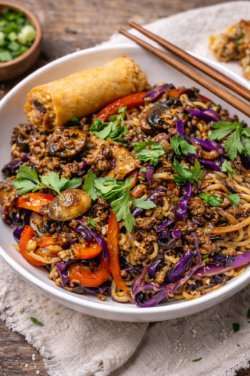 Plated beef ramen noodle dish garnished with fresh cilantro, served alongside an egg roll on a white plate