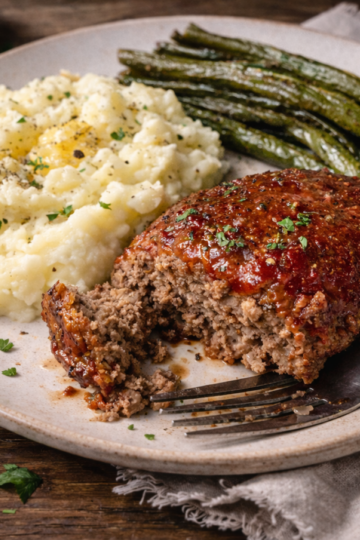 A plated dinner featuring a slice of meatloaf with ketchup glaze, creamy mashed potatoes, and roasted green beans, with a fork on the plate.