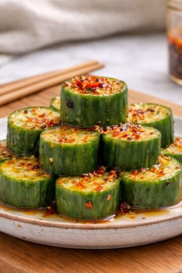 Asian cucumber salad served on a white ceramic plate with thick cucumber rounds arranged in a pyramid, topped with red chili flakes and drizzled with a soy-based sauce. Chopsticks rest nearby on a wooden board.