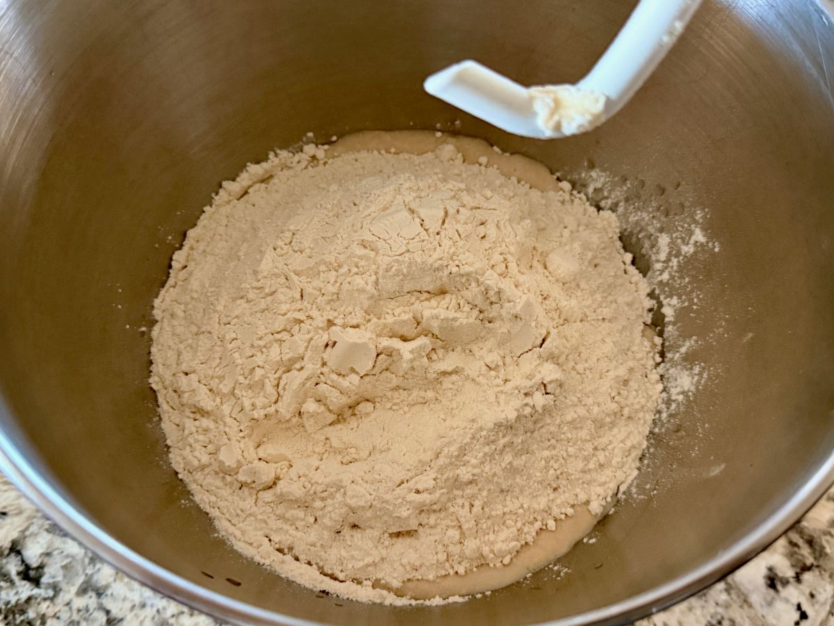 Flour being added to a stand mixer bowl while mixing dough for homemade onion rolls.