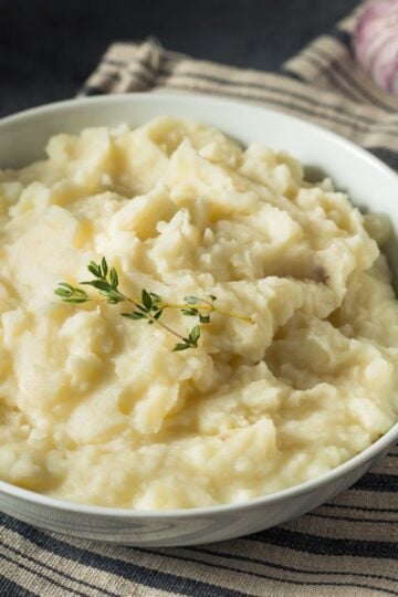 Finished bowl of mashed potatoes on a striped dish towel with a spoon and a bulb of garlic in the background