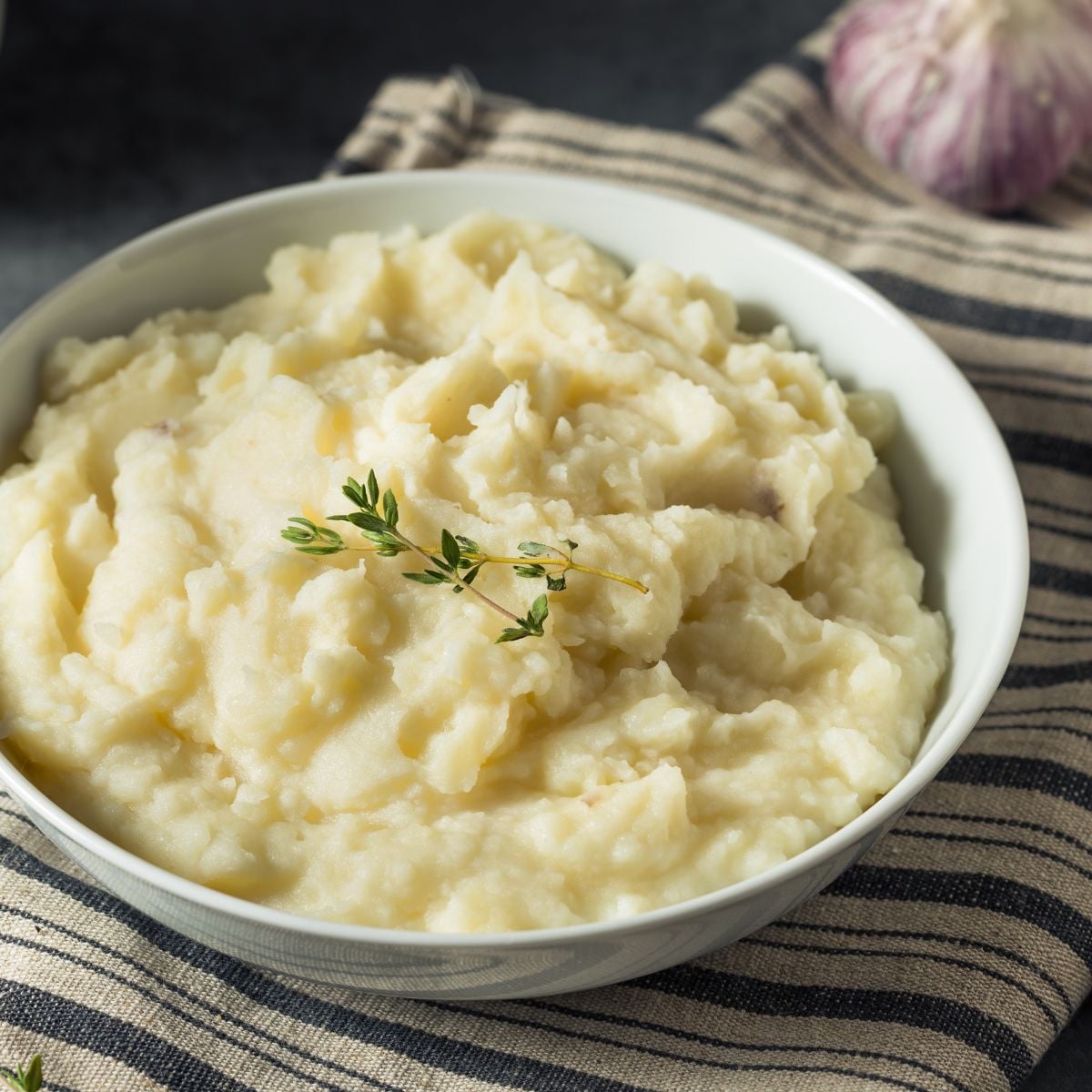 Finished bowl of mashed potatoes on a striped dish towel with a spoon and a bulb of garlic in the background