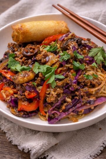 Plated beef ramen noodle dish garnished with fresh cilantro, served alongside an egg roll on a white plate