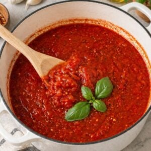 Close-up of thick homemade marinara sauce in a white Dutch oven with a wooden spoon and fresh basil on top