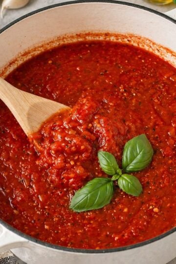 Close-up of thick homemade marinara sauce in a white Dutch oven with a wooden spoon and fresh basil on top