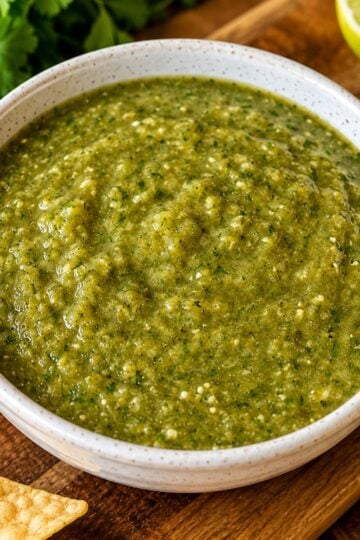 Homemade roasted salsa verde in a white ceramic bowl served with tortilla chips and fresh lime wedges.